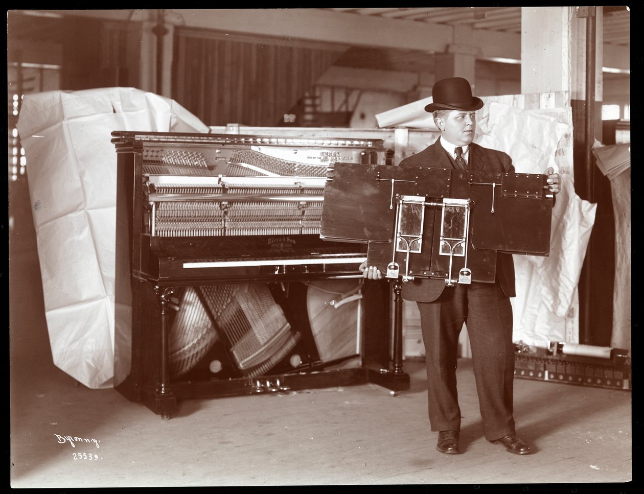 Ett piano av Ricca and Son Piano Co. med en man bredvid som håller upp en del av pianoverken, 1907 (silvergelatintryck) av Byron Company