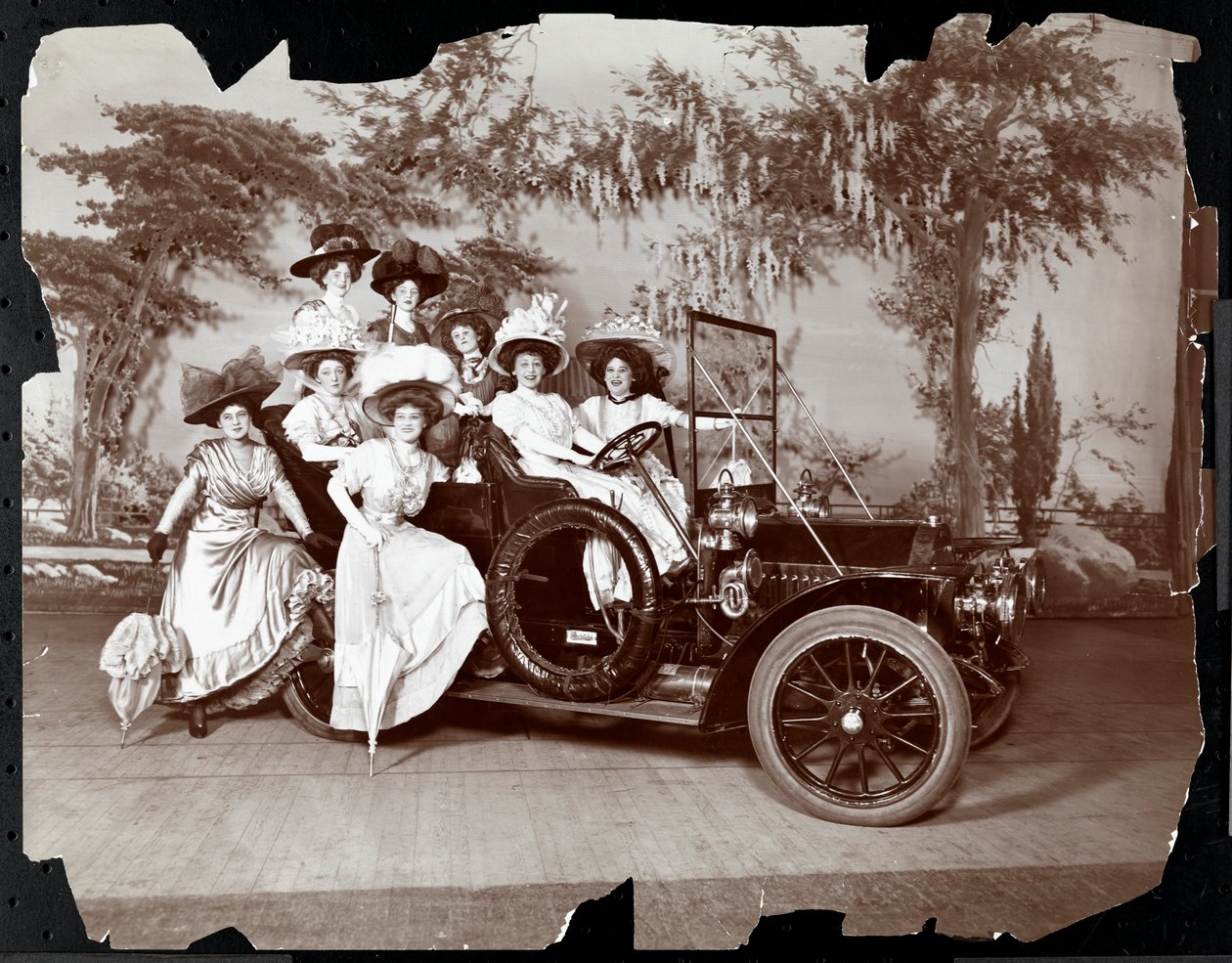 Åtta kostymklädda kvinnor poserar i och runt en bil på en scen med ett landskap i bakgrunden, troligen i samband med föreställningen "The Vanderbilt Cup", 1906 (silvergelatintryck). av Byron Company