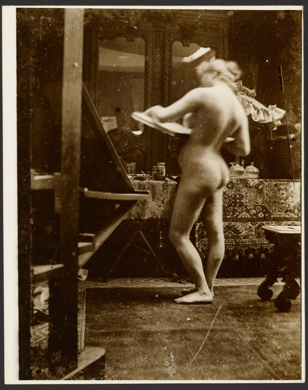 [Kvinnlig nakenmodell står i studio och håller i en skissbok] av Alphonse Maria Mucha