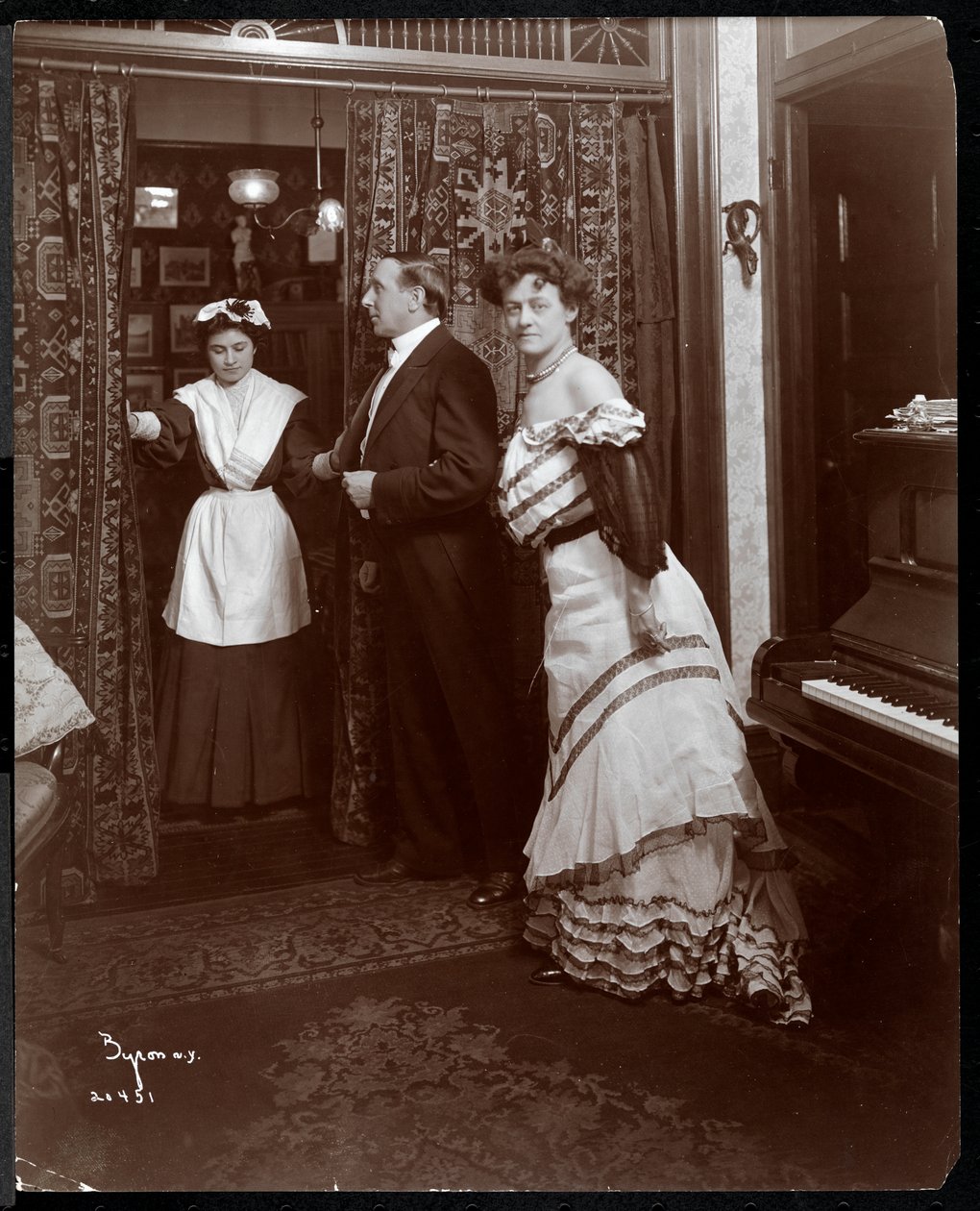En formellt klädd man och kvinna och vad som ser ut att vara en hemtjänst i ett rum med ett piano, 1905 (silvergelatintryck) av Byron Company