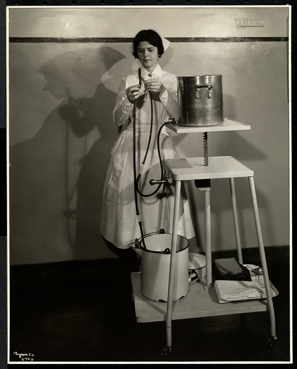 En sjuksköterska som demonstrerar en speciell apparat med rör och förråds- och avfallshinkar på Sloan Maternity Hospital, Broadway och 168th Street, New York, 1932 (silvergelatintryck) av Byron Company