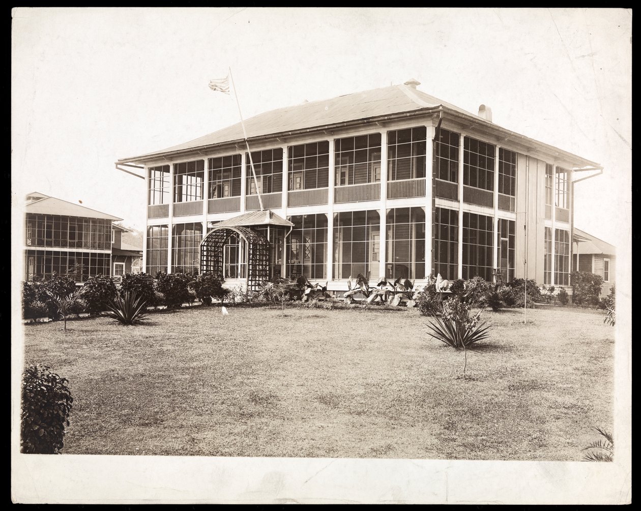 Ett tvåvåningshus omgivet av verandor, med en amerikansk flagga vajande; i Panama under byggandet av kanalen, 1912 (silvergelatintryck) av Byron Company