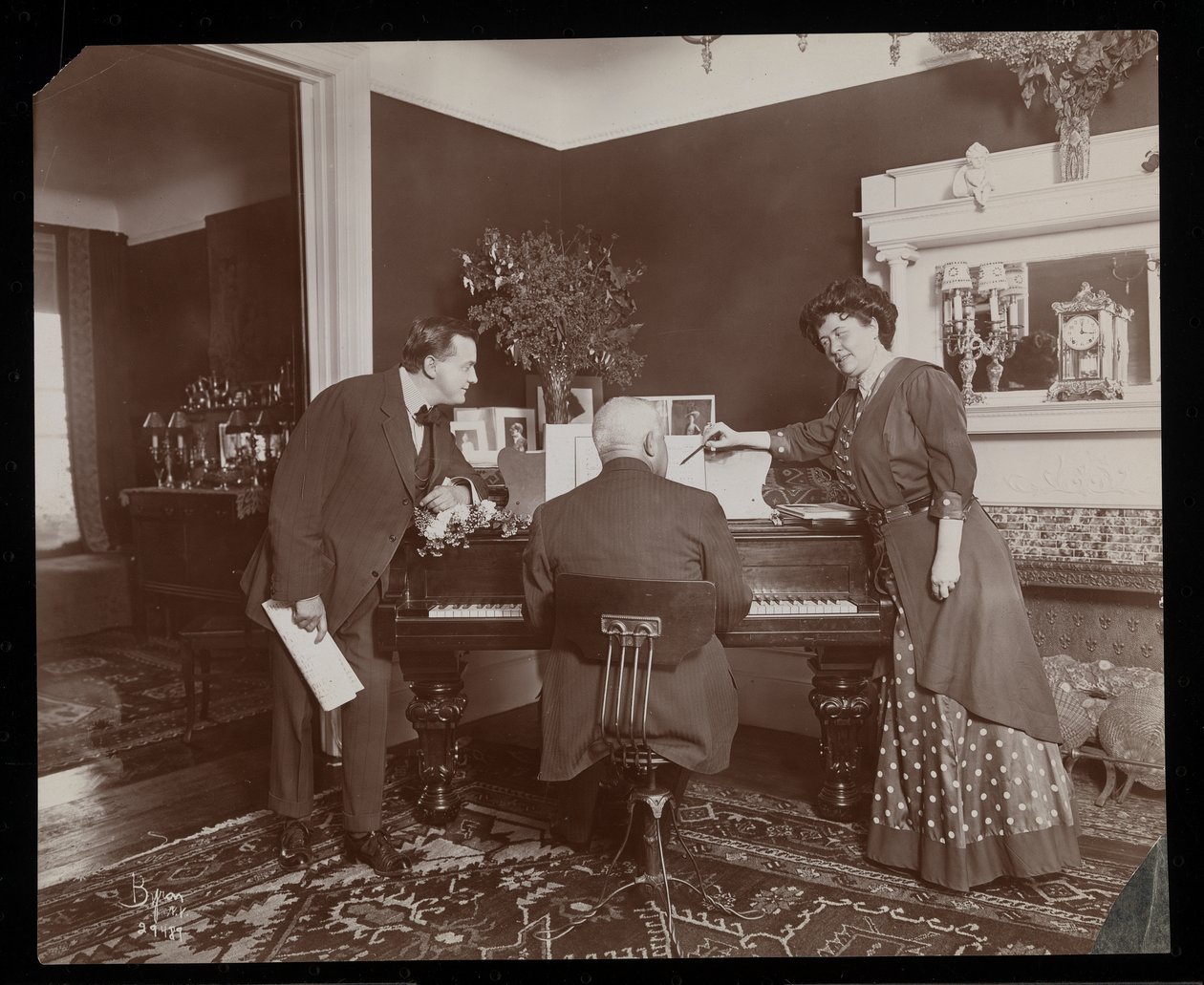 Clara De Regand pekar på ett partitur medan en man spelar piano, 1910 (silvergelatintryck) av Byron Company