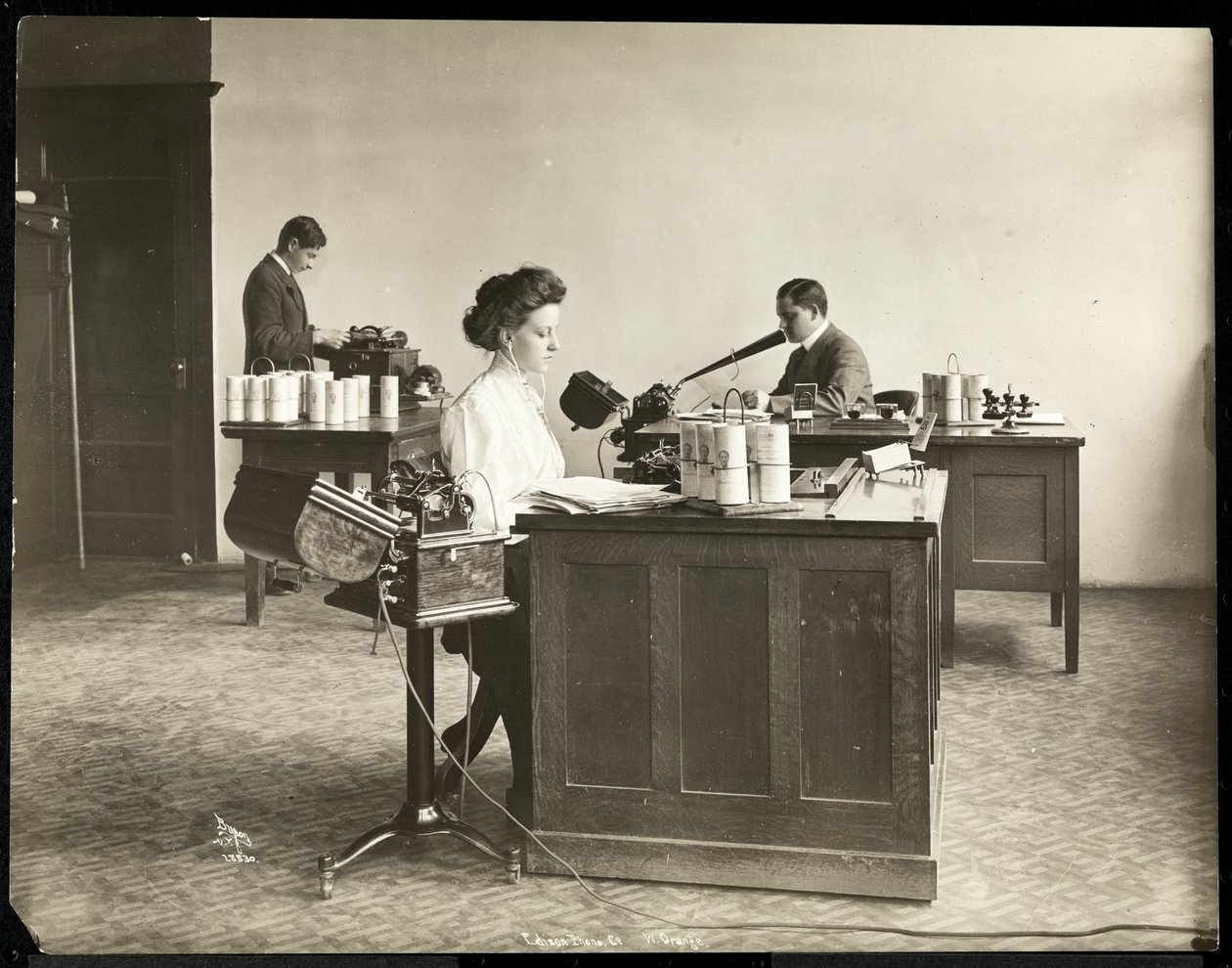 Interiörvy av en kvinna som skriver från en dikteringsmaskin och en man som dikterar i en annan på Edison Phonograph Co., West Orange, New Jersey, 1903 (silvergelatintryck) av Byron Company