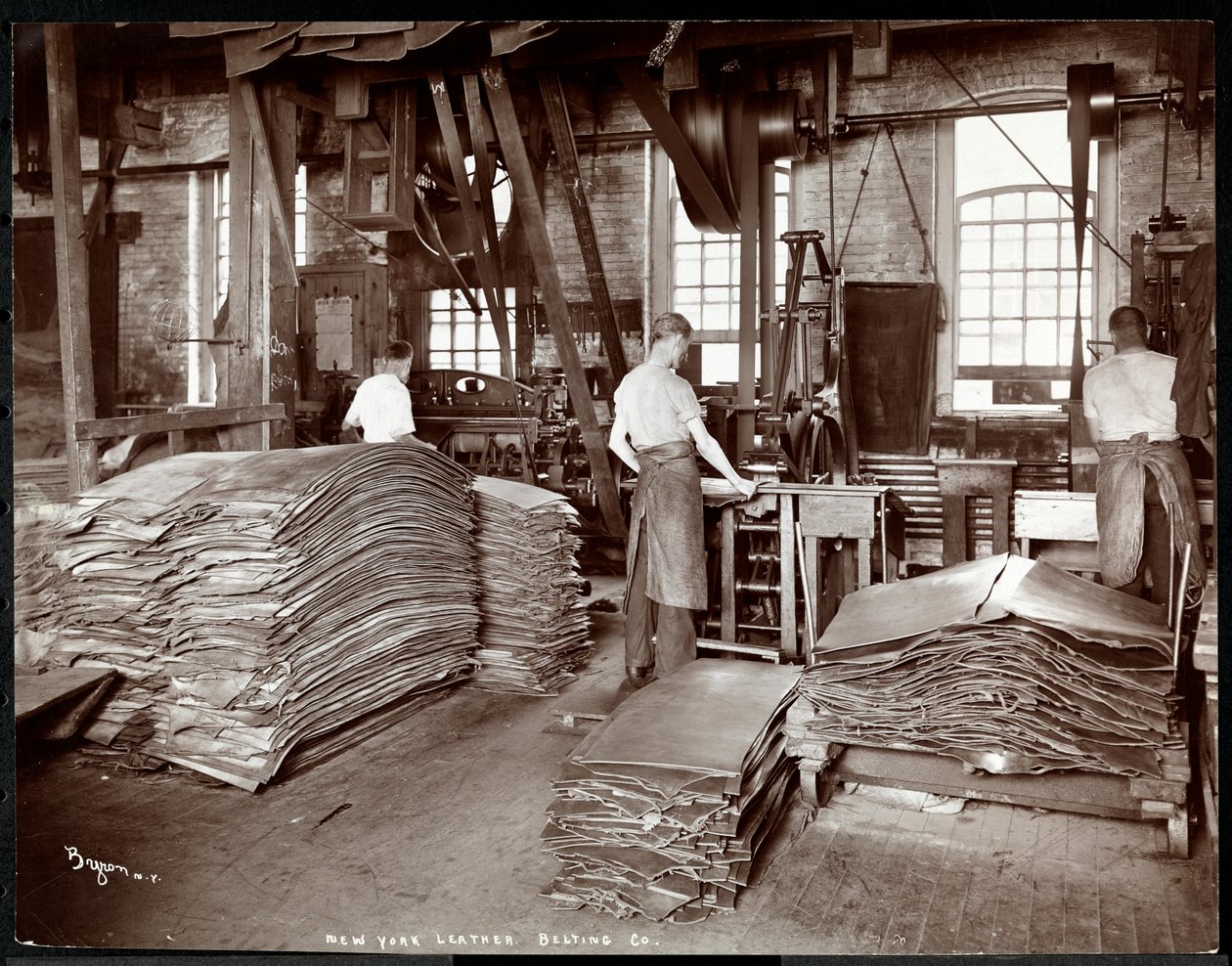 Interiör av tre män som arbetar med läder och tung utrustning på New York Leather Belting Co. i New York, 1905 (silvergelatintryck). av Byron Company
