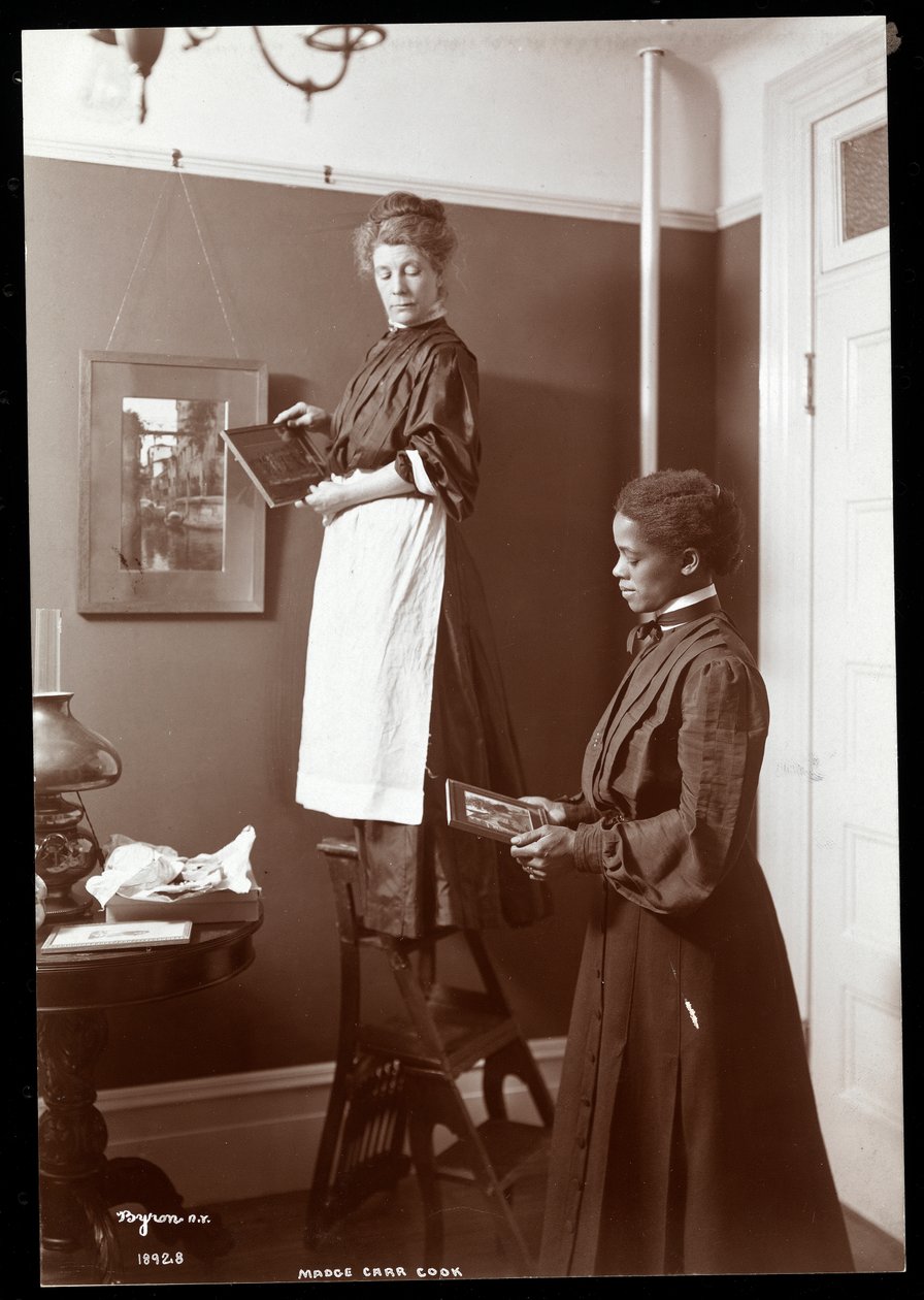 Madge Carr Cook hänger tavlor tillsammans med sin piga, ca 1904 (silvergelatintryck) av Byron Company