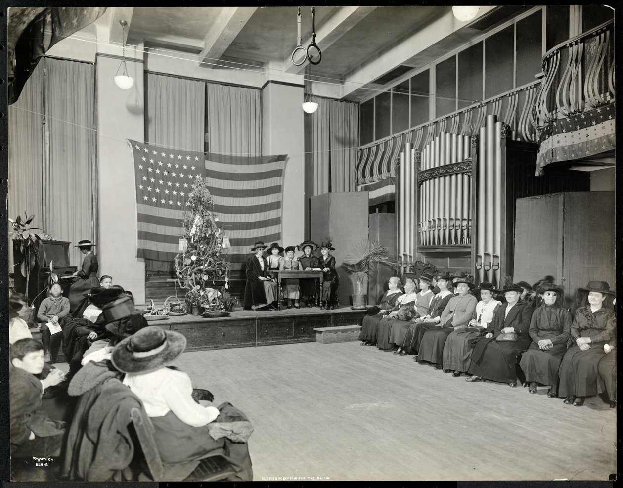 Människor i ett auditorium på New York Association for the Blind med julgran och personer som möjligen framför en sketch på scen med orgel, 1920 (silvergelatintryck) av Byron Company