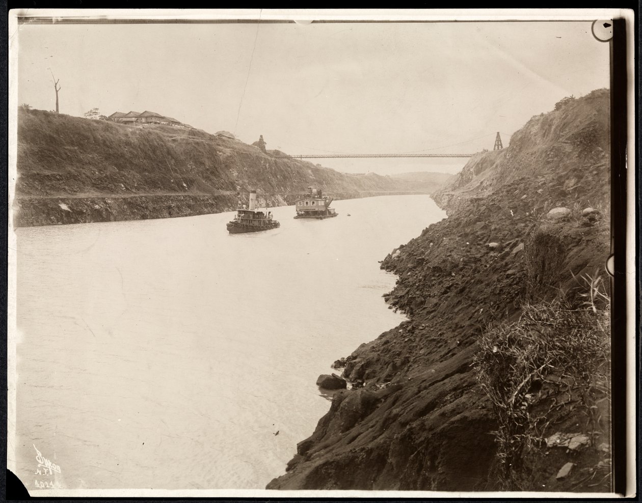 Vy över två båtar i Panamakanalen under dess konstruktion med en hängbro bortom, 1913 (silvergelatintryck) av Byron Company