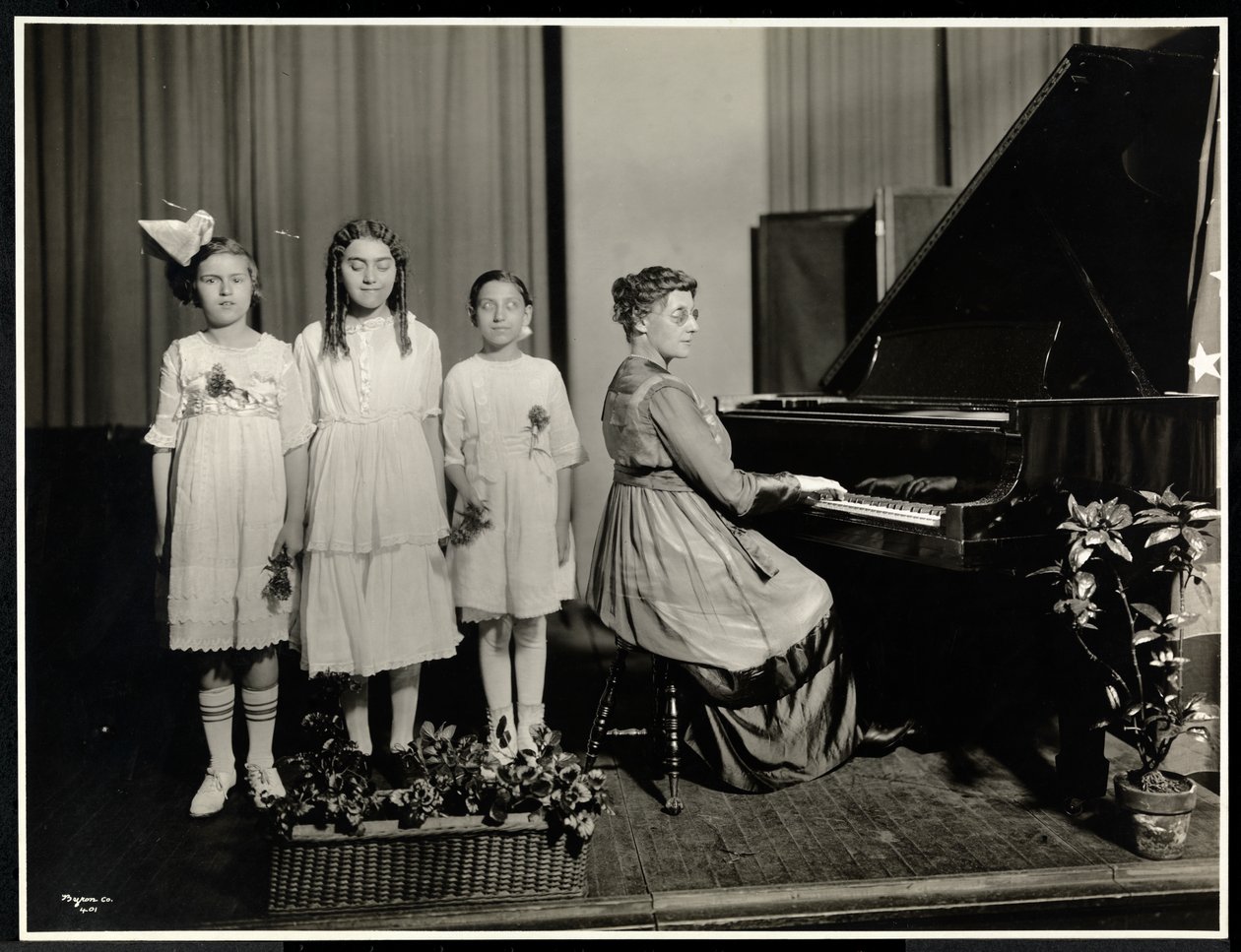 En kvinna spelar piano medan tre flickor sjunger på New York Association for the Blind, 111 East 59th Street, New York, ca 1917 (silvergelatinprint). av Byron Company