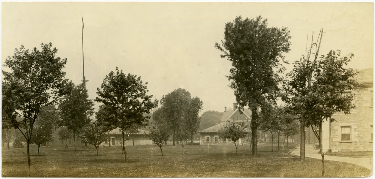 Horizontal, sepia photograph showing a group of brick buildings bordering on an open green space with trees scattered throughout. Some of the brick buildings appear to be houses av Carl Michel