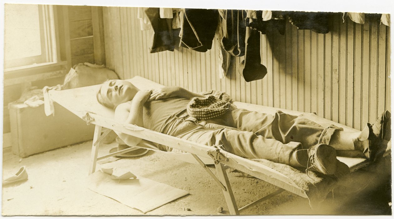 Horizontal, sepia photograph showing a man sleeping on a cot in a wood paneled building. Various articles of clothing can be seen hanging on the wall, and suticases can be seen sitting on the ground in the corner of the room av Carl Michel