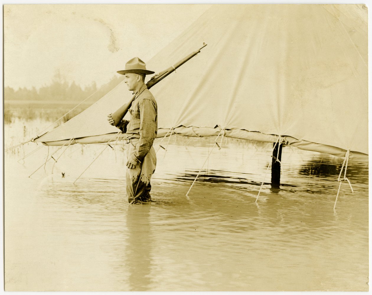 Horizontal, sepia photograph showing a uniformed soldier standing guard in a flooded field. The soldier is standing next to a tent, which has its sides rolled up and secured above the water line av Carl Michel