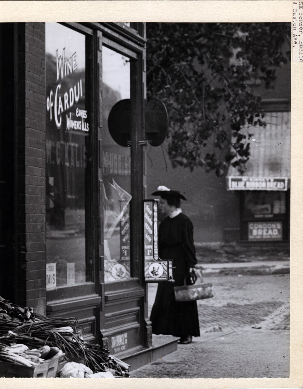 Vertical, black and white photograph showing a woman holding a basket while standing in front of the W. C. Kalis Drugstore entrance at 4878 Easton Avenue av Charles Clement Holt