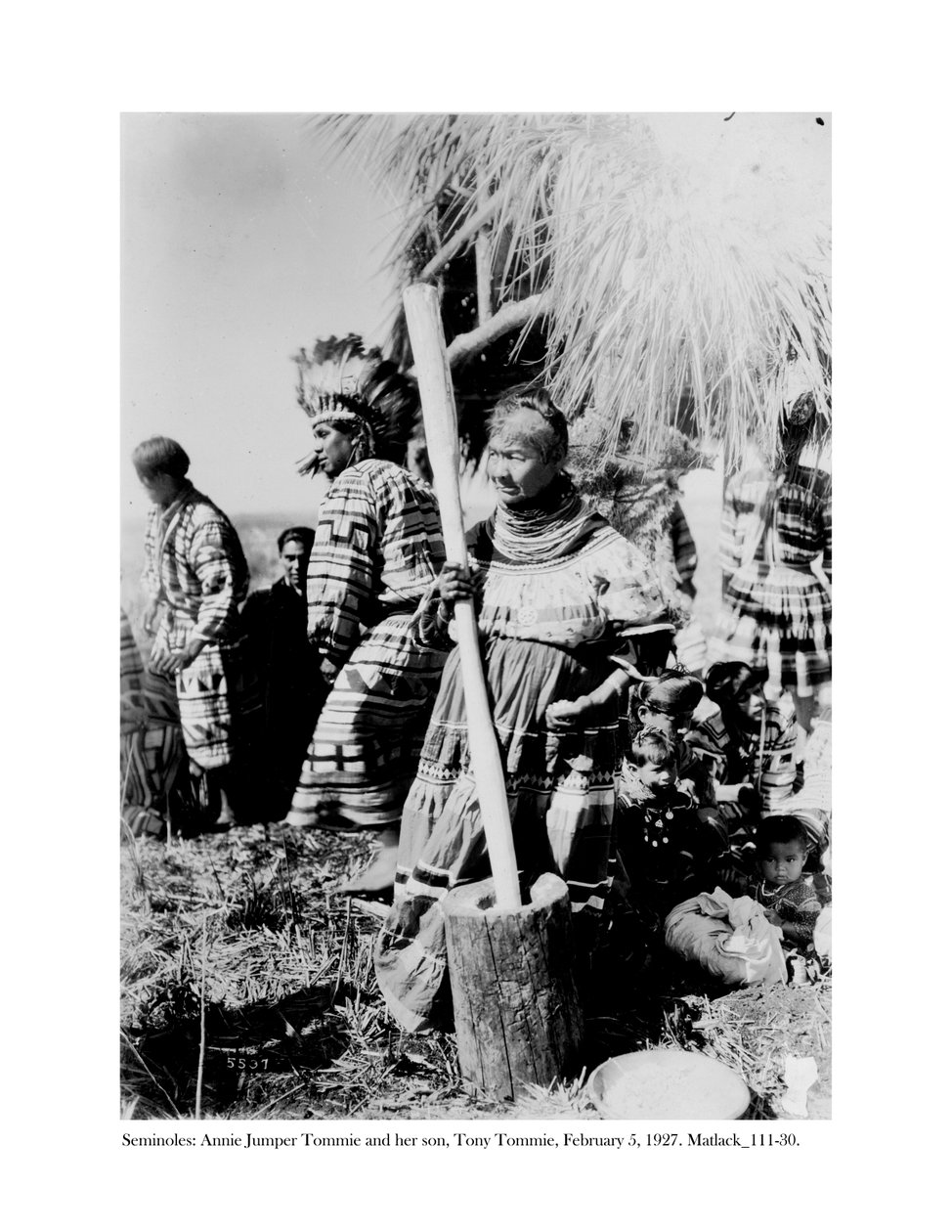 Annie Jumper Tommie använder en mortel och mortelstöt för att göra majsmjöl, och hennes son, Tony Tommie, bär en örnfjäderhuva, 5 februari 1927 av Claude C. Matlack