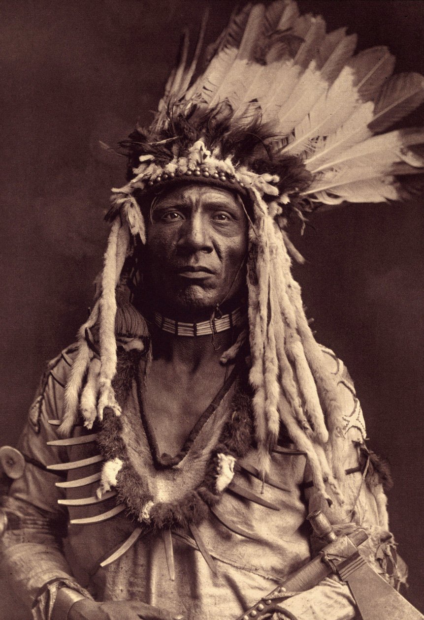 Vesselsvans (Apohsuyis). Denna indiska Piegan bär den berömda krigshäcken i örnfjädrar och vesslingskinn, en mockatunika, två halsband, ett i ben och det andra i grizzlyklor, och en tomahawkpipa. Foto tagen från volym 6 av det publicerade uppslagsverket av Edward Sheriff Curtis