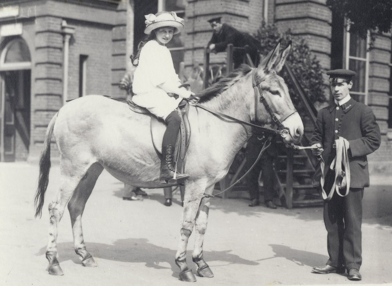 En ung dam som rider på en rumpa som leds av en målvakt medan en annan ser på, London Zoo maj 1914 av Frederick William Bond