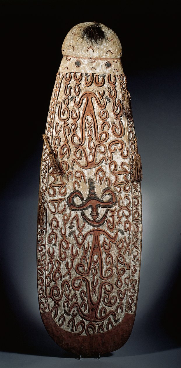 Oval Asmat-sköld, snidad i relief med rullande ornament som innehåller mycket stiliserade figurer, flätade tofsar till bården (trä, raffia och fjädrar) av Guinean New