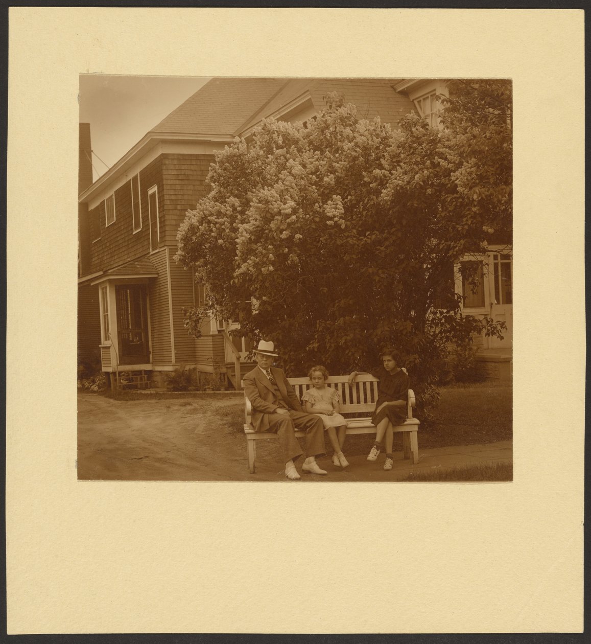 [Man och unga flickor på en bänk framför ett hus] av Louis Fleckenstein