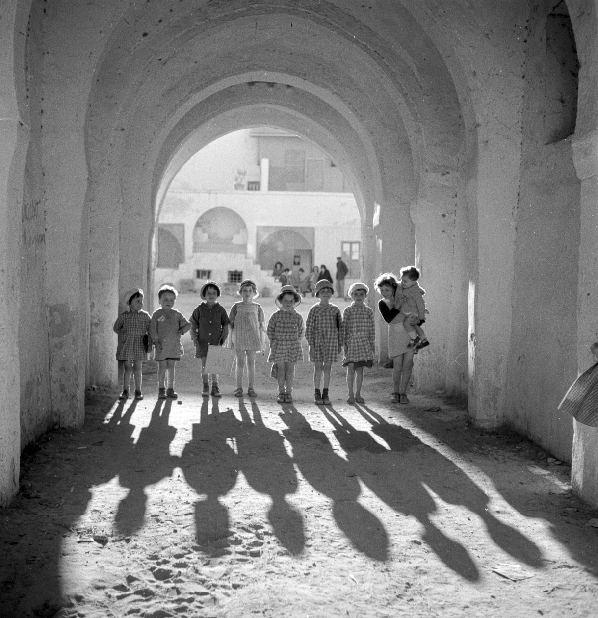 Europeiska barn i skoluniform under båge med skuggor. av Maynard Owen Williams