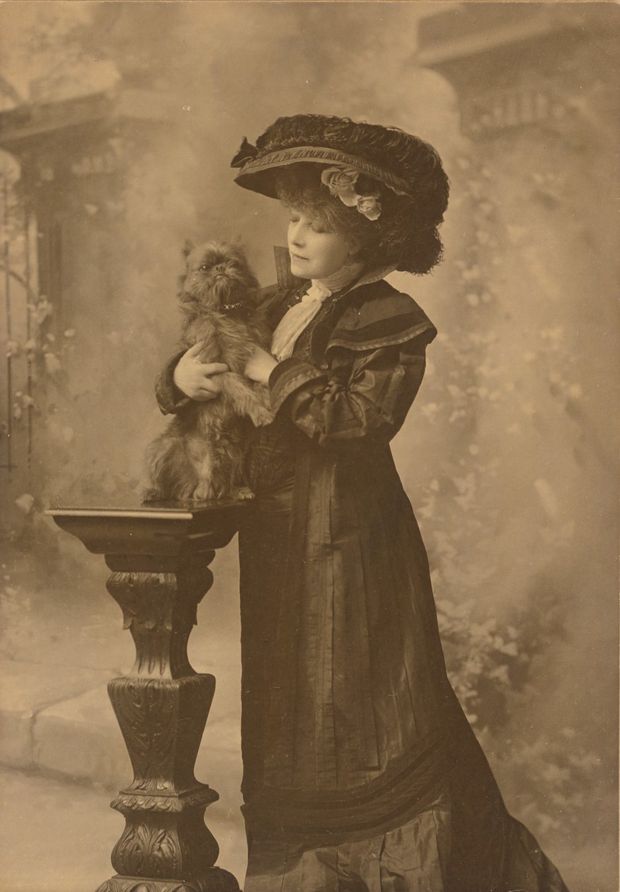 [Sarah Bernhardt står i en studiomiljö och håller i en liten svart hund på en utsmyckad, snidad träpiedestal] av Theodore C. Marceau