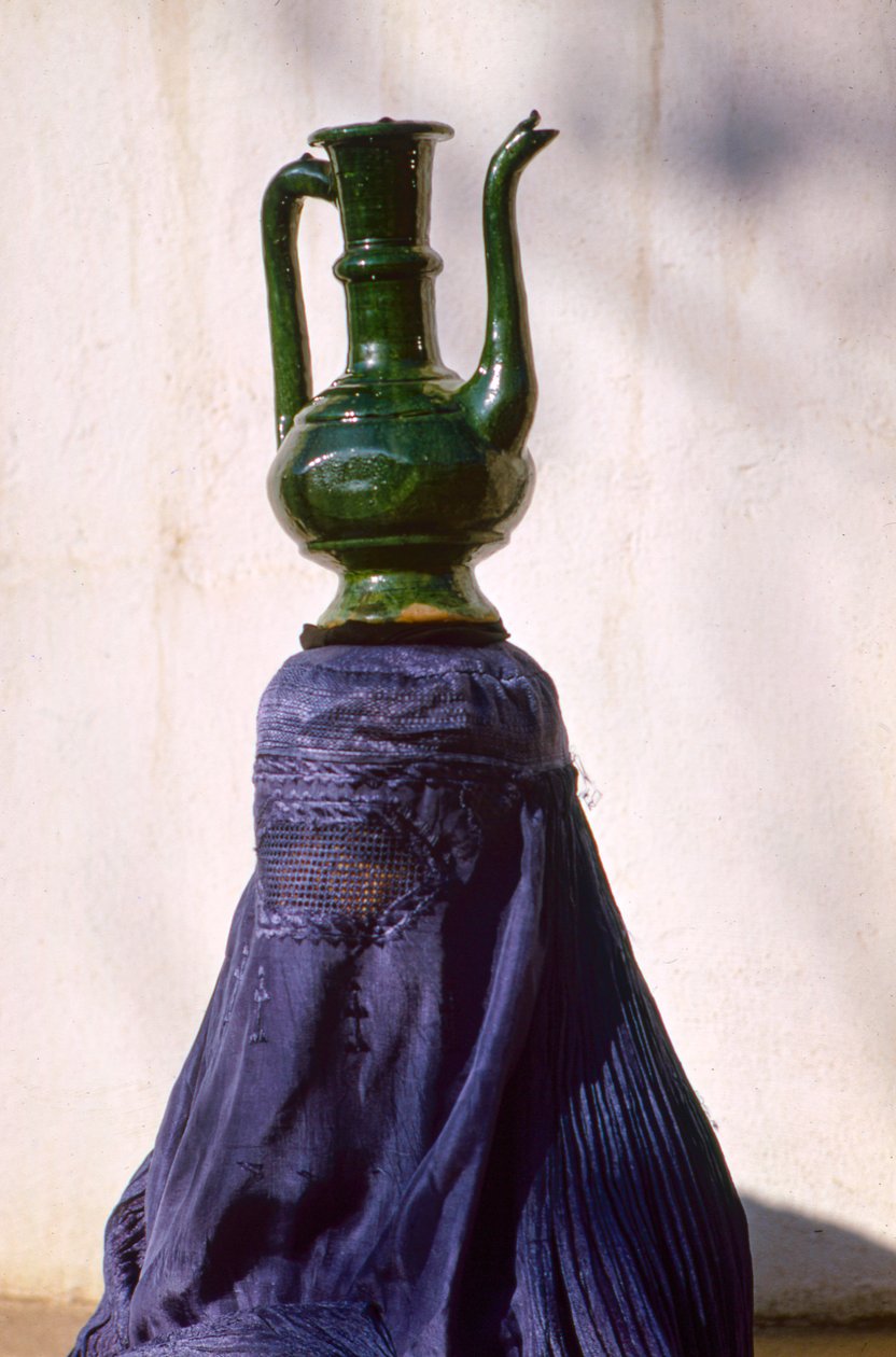 Kvinna i Kabul som är skyddad från insyn av en chadri aka chador-slöja i siden bär en kanna från en... av Thomas J. Abercrombie