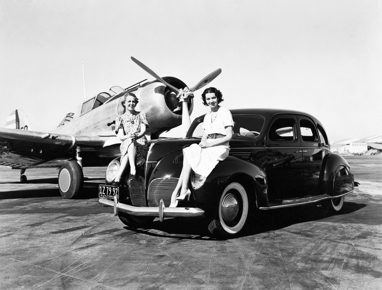 Kalifornien: 1939 Två kvinnor sitter på stänkskärmarna på en Lincoln Zephyr sedan 1939 på en flygplats med ett enmotorigt plan parkerat bakom dem (s/v foto) av Unbekannt