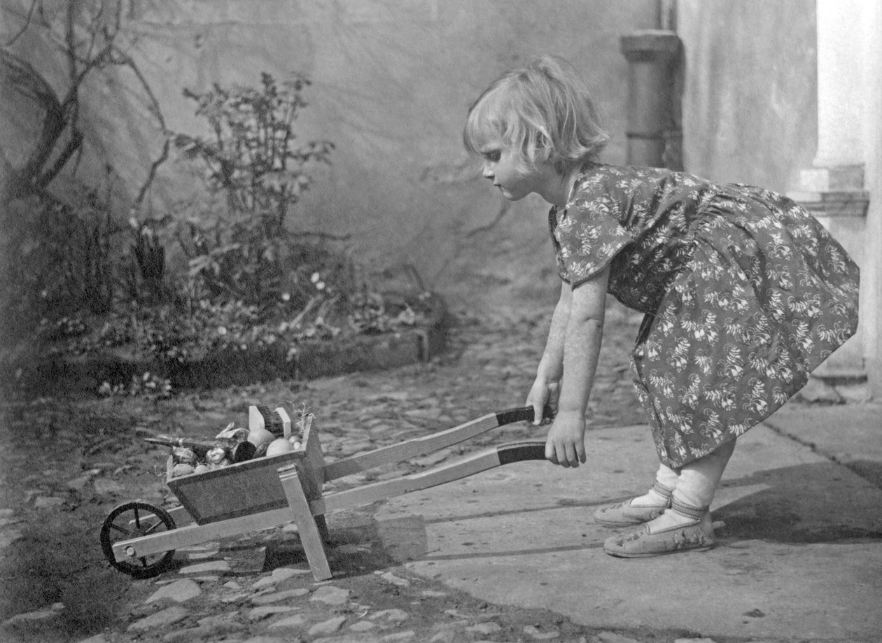 Liten flicka som leker med en skottkärra. Fotografering omkring 1931 av Unbekannt