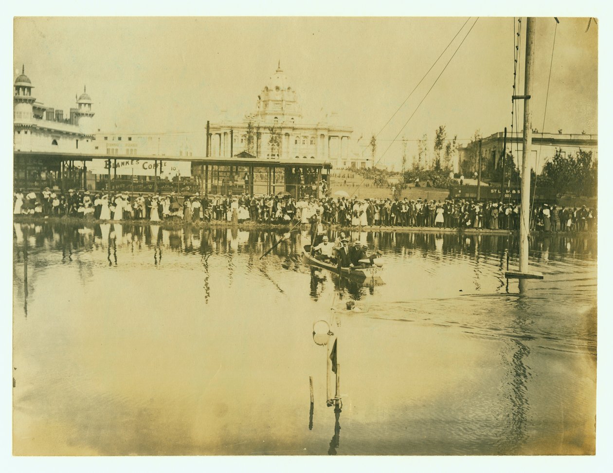 Horizontal photograph of a swimmer crossing a finish line. Men in a boat watch as well as crowds of people on the shore av Unknown