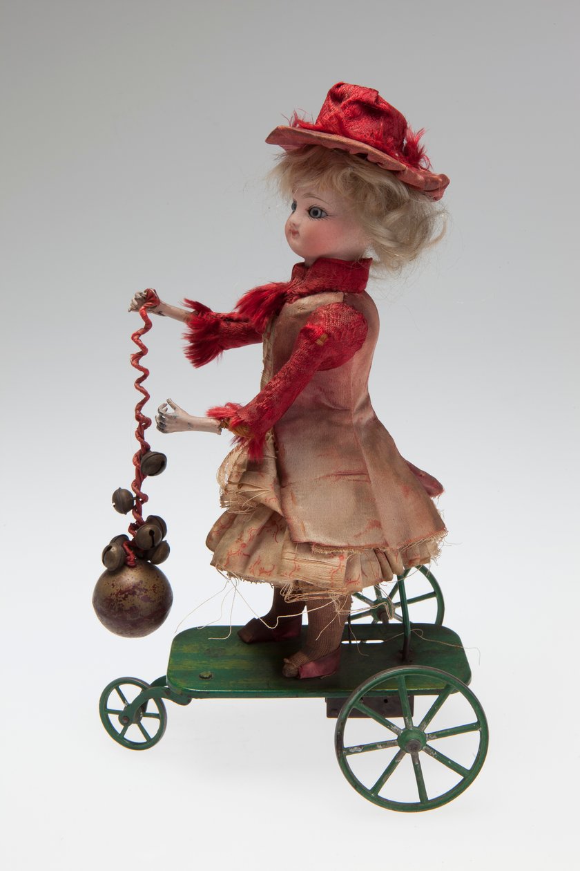 Mechanical clockwork doll on metal wheeled platform that rings a bell and turns her head as the mechanism turns av Unknown