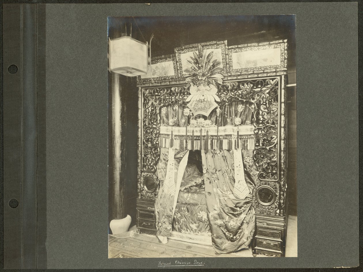Vertikalt, sepia fotografi som visar en baldakinsäng med en utförligt snidad sängstolpe i trä och draperier och överdrag av silke. av Unknown