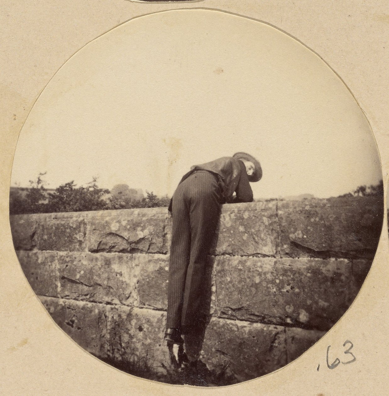 [Man i hatt böjd över en stenmur] av Unknown artist