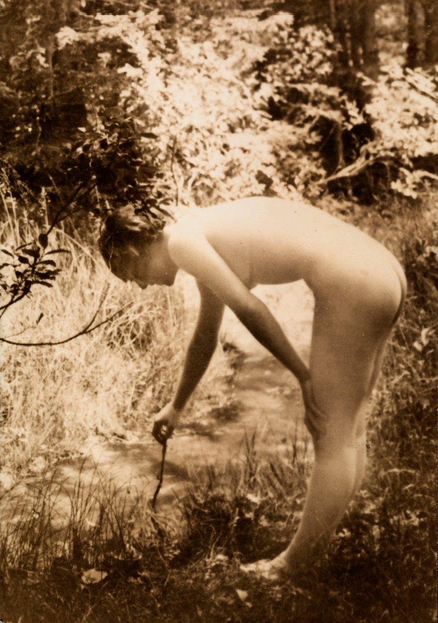 En ung kvinna, naken i skogen, ses i profil när hon böjer sig fram för att lägga en gren i en bäck. av Unknown photographer