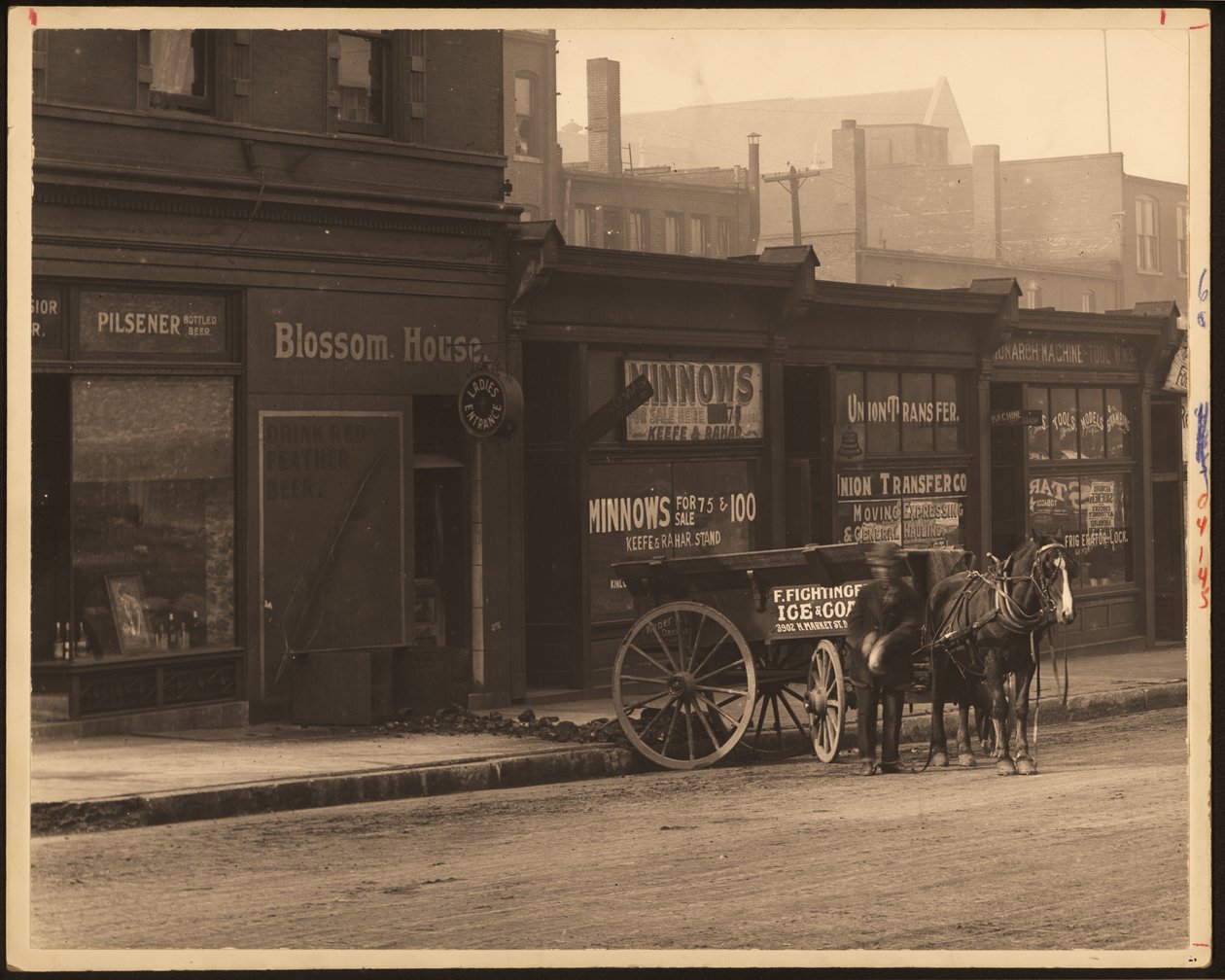 Horizontal, black and white photograph of a row of commercial buildings and a horse-drawn wagon parked against the sidewalk av William G. Swekosky