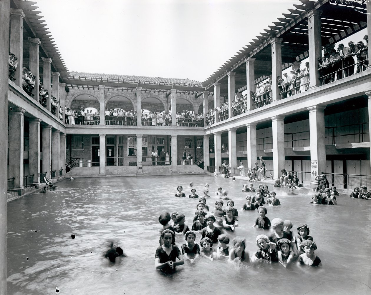 Horizontal, black and white photograph of a swimming pool with two story balcony supported by columns. Onlookers watch the swimmers from the second level balcony av William G. Swekosky