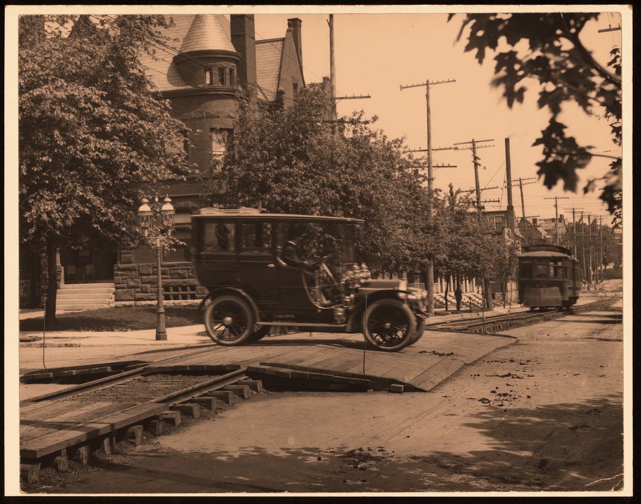 Horizontal, black and white photograph of an automobile crossing over trolley tracks as a trolley moves down the track av William G. Swekosky