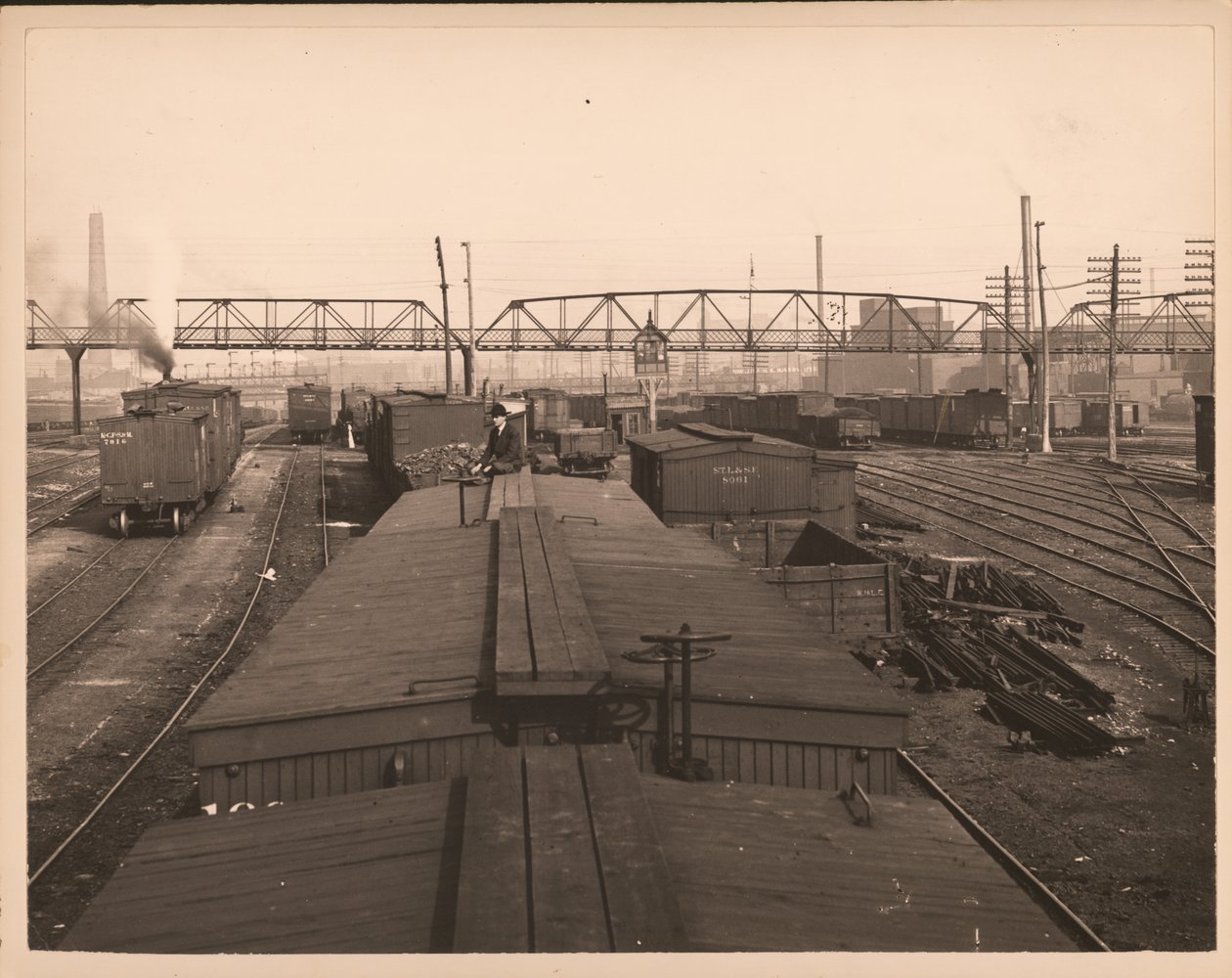 Horizontal, black and white photograph of rail cars in a rail yard. A man sits on top of one of the cars av William G. Swekosky
