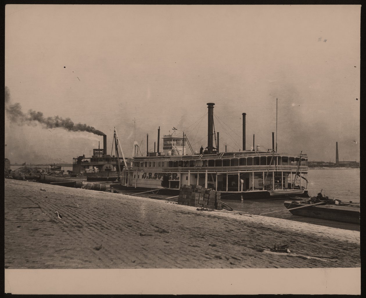 Horizontal, black and white photograph of the steamboat C.R. Suter. Another steamboat is parked behind it and crates are piled on the shore av William G. Swekosky