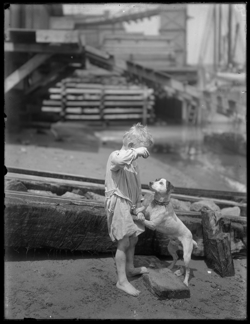 Barfotapojke, möjligen William Gray Hassler, leker med hund på stranden av en flod eller sjö, New York, ca 1910 av William Davis Hassler