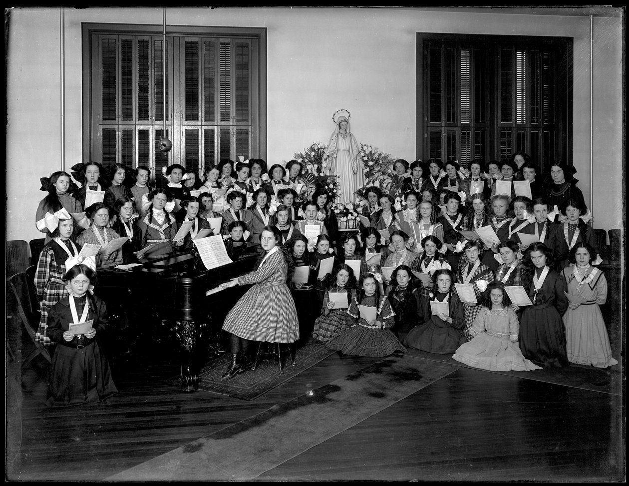 Gruppporträtt av en flickkörklass samlad kring ett piano som håller noter, troligen på det romersk-katolska föräldrahemshuset i Kingsbridge, Bronx, ca 1911 av William Davis Hassler