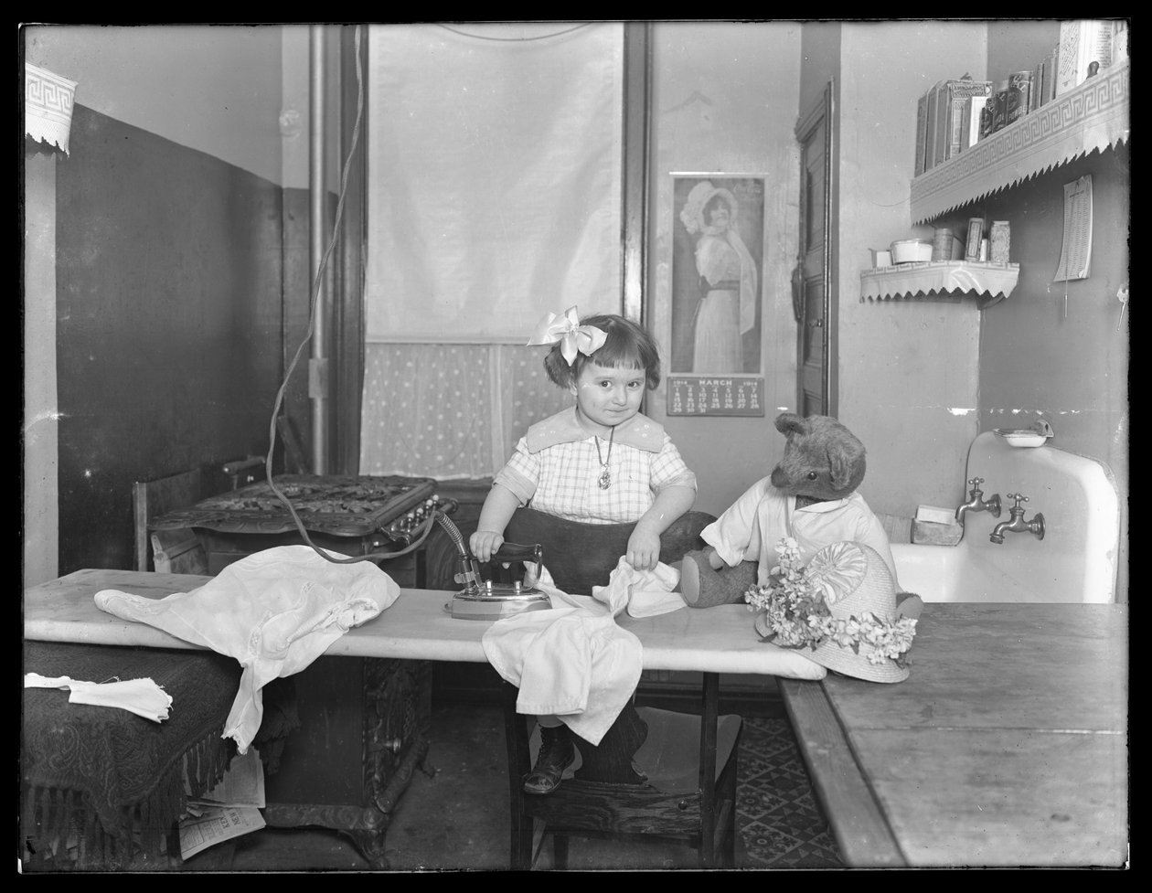 Oidentifierad liten flicka som visar hur man använder ett elektriskt strykjärn, ca 1913-14 av William Davis Hassler