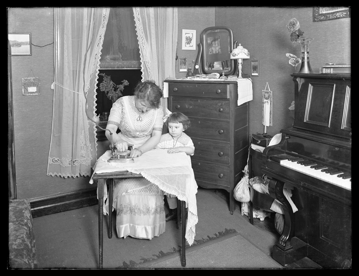 Oidentifierad kvinna som visar användningen av ett elektriskt strykjärn, bevakad av en liten flicka, ca 1913-14 av William Davis Hassler
