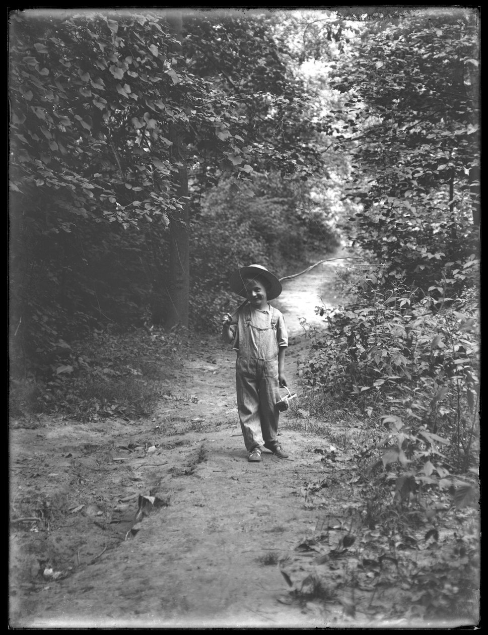 William Grey Hassler i stråhatt och overall, bärande fiskespö och korg, går längs en stig ensam i skogen, 20 augusti 1911 av William Davis Hassler