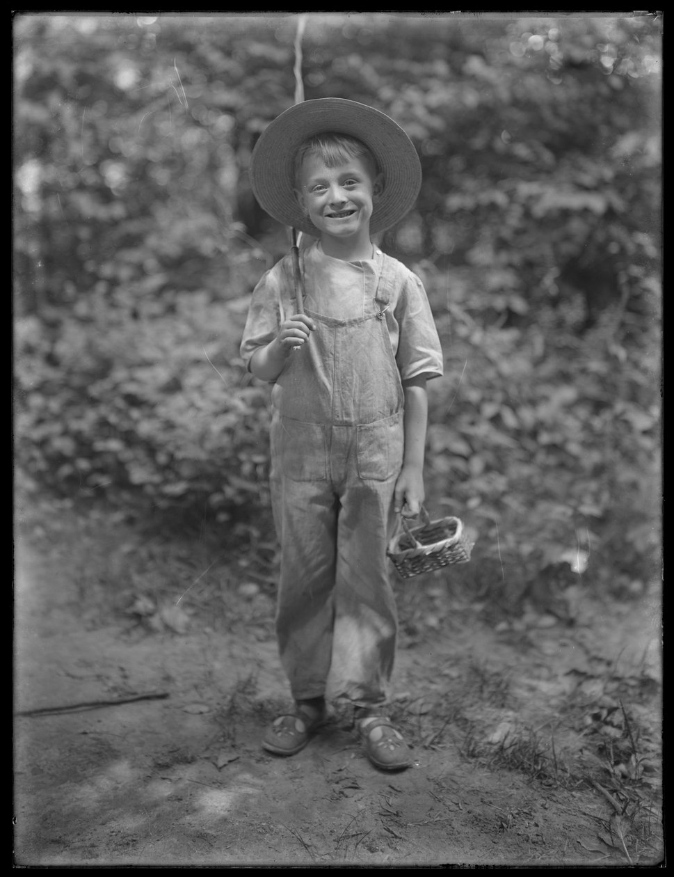 William Grey Hassler i stråhatt och overall, bärande fiskespö och korg i skogen, 20 augusti 1911 av William Davis Hassler