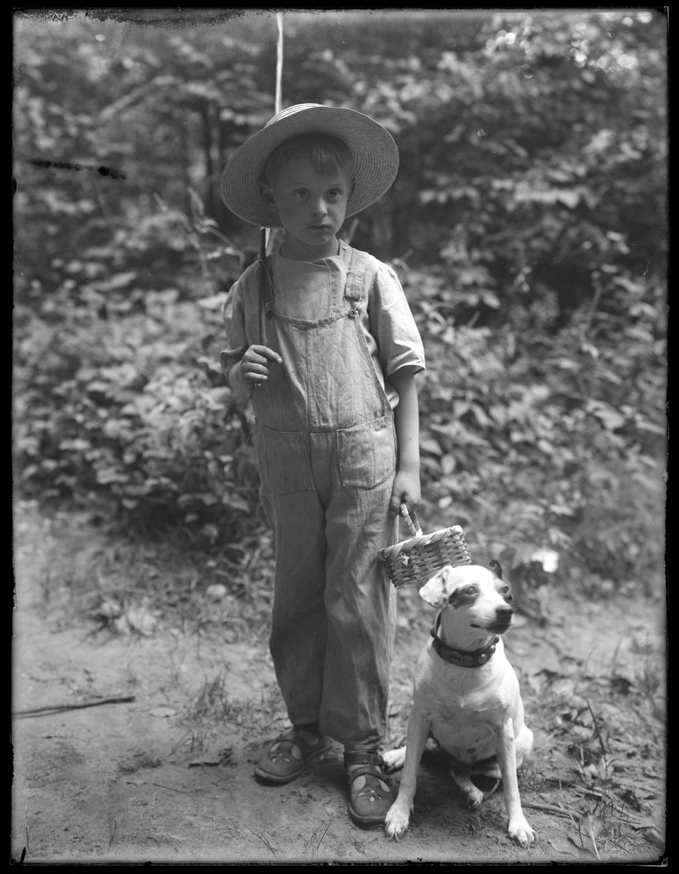 William Gray Hassler i halmhatt och overall, med fiskespö och korg, poserar i skogen med en liten hund (Bessie), 20 augusti 1911. av William Davis Hassler