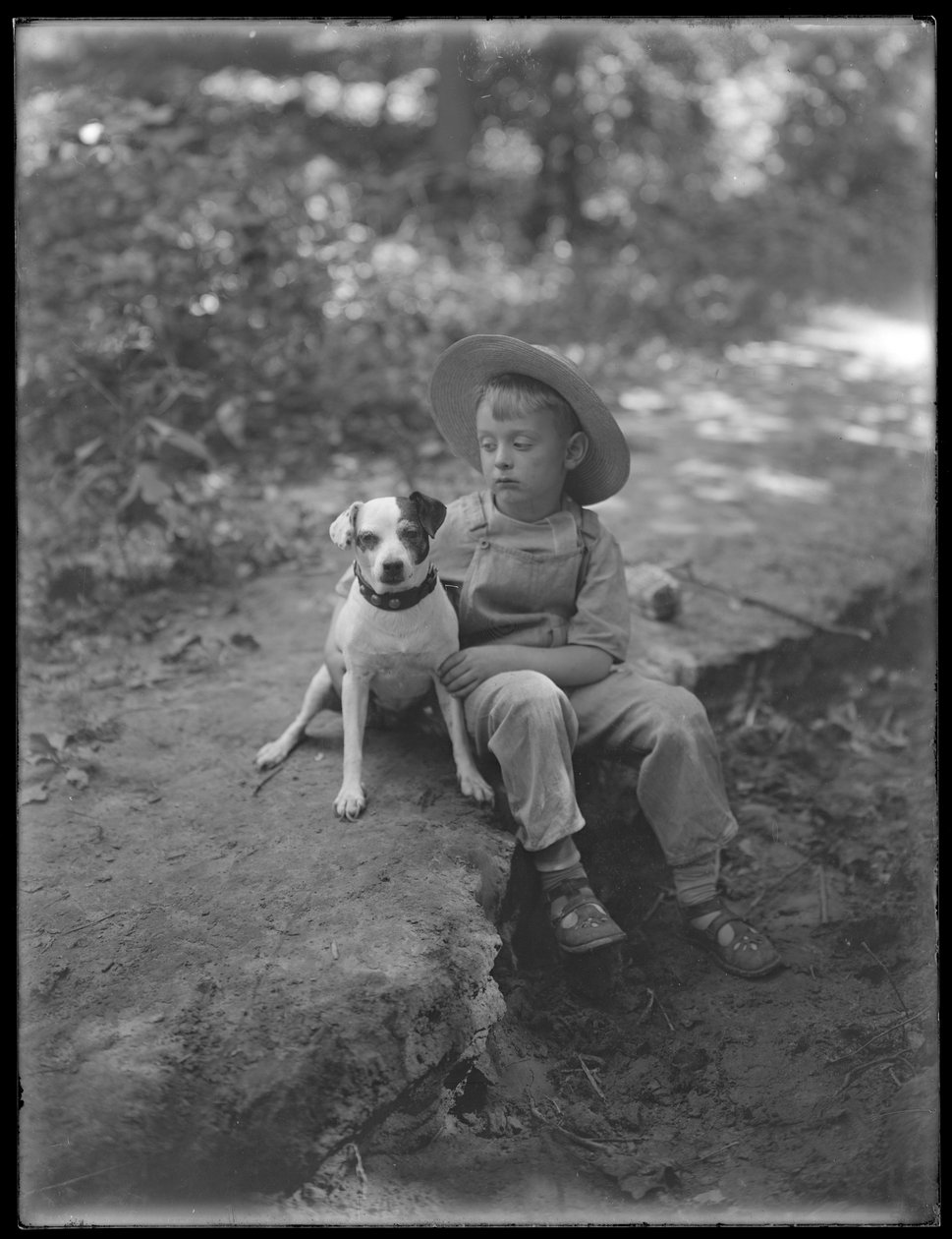 William Grey Hassler i stråhatt och overall, poserade sittande i skogen med en liten hund (Bessie), 20 augusti 1911 av William Davis Hassler