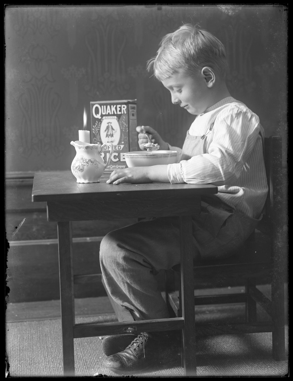 William Gray Hassler sittande vid ett litet bord och äter en skål med Quaker puffade risflingor, ca 1912 av William Davis Hassler