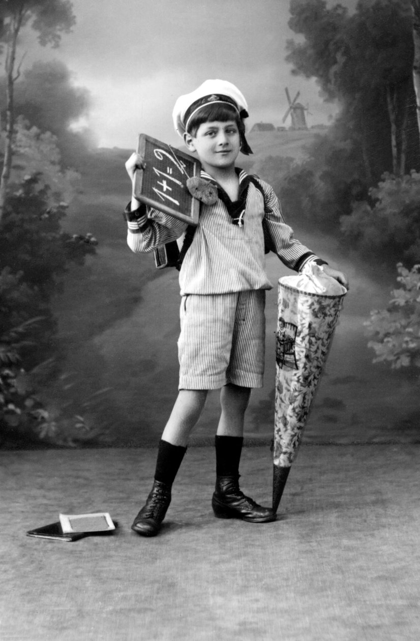 Första dagen på skolan. En ung pojke i uniform poserar med en tavla som visar tillägget 1+1 = 2 och en överraskningspåse. Fotografering omkring 1910. av Ferdinand Georg Waldmuller