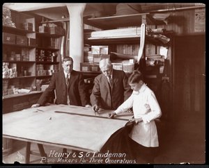 Porträtt av Henry och S.G. Lindeman med deras son eller barnbarn stående över butiksteckningar för ett piano, 1910 (silvergelatintryck)
