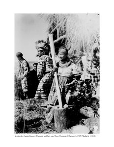Annie Jumper Tommie använder en mortel och mortelstöt för att göra majsmjöl, och hennes son, Tony Tommie, bär en örnfjäderhuva, 5 februari 1927