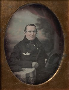 Man som bär kappa och handskar, håller hatt, sitter framför målad utomhusbakgrund, 1840-talet. av Samuel Broadbent Jr..